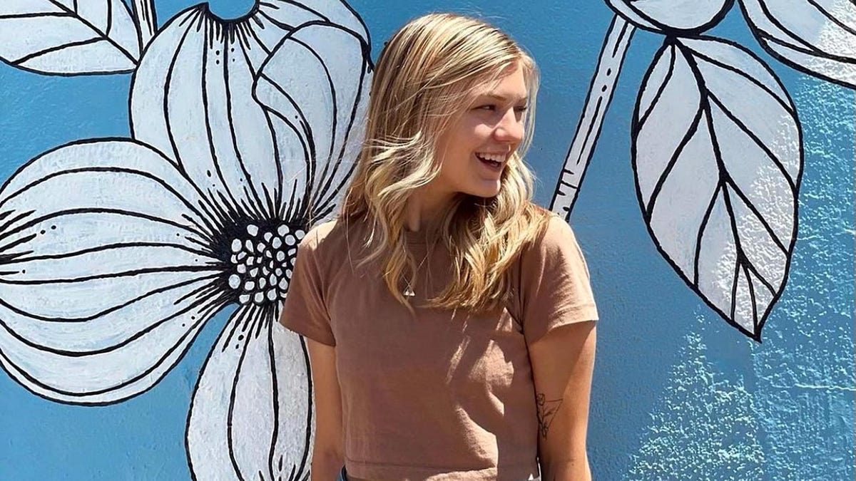 Gabby Petito poses in front of a blue and white mural of flowers while looking over her shoulder