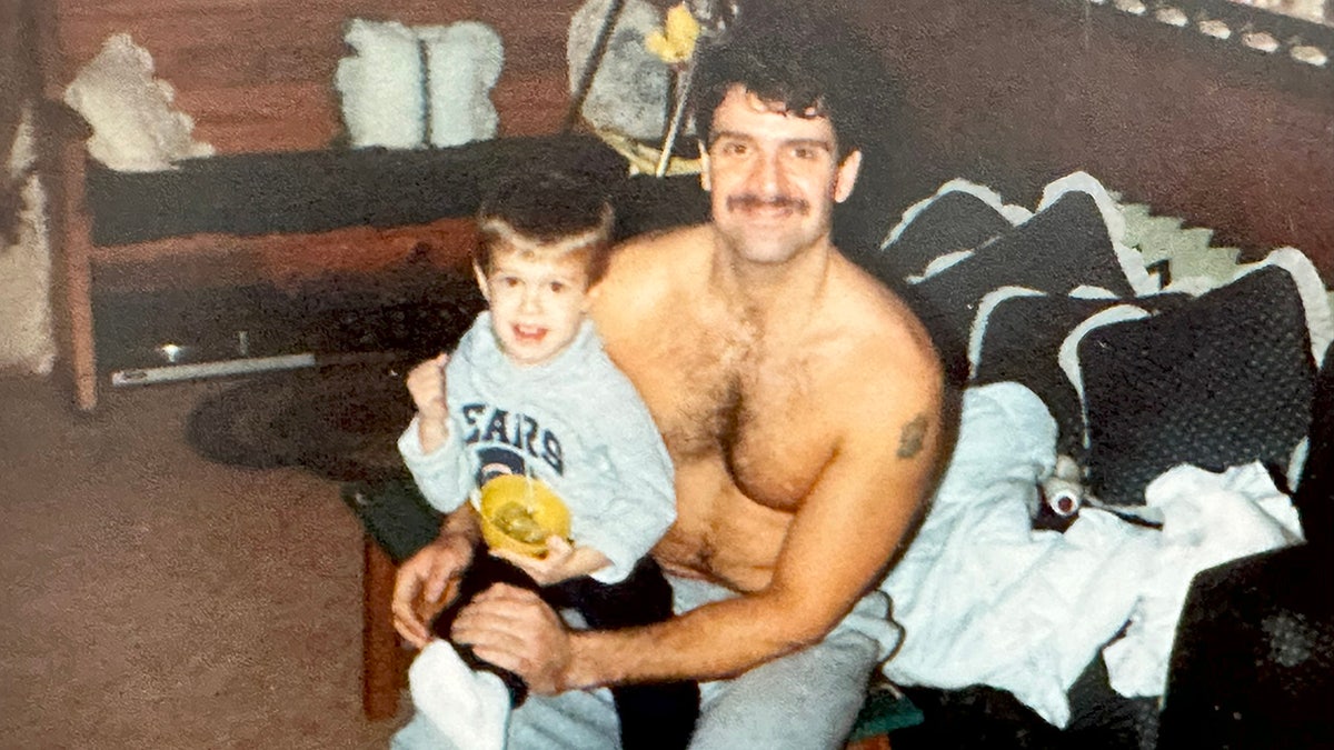 Ken Tekiela shirtless and wearing grey sweatpants holding his son Kyle.