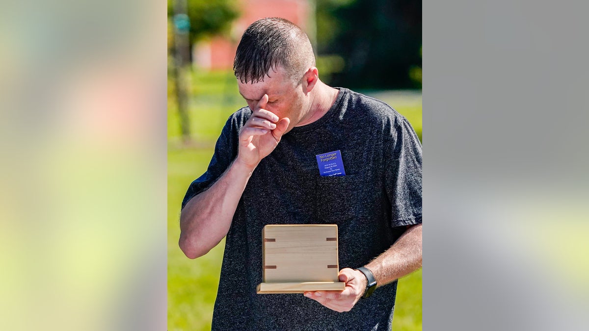 Eric Pranger wearing a navy shirt holding a wooden box and wiping a tear outdoors.