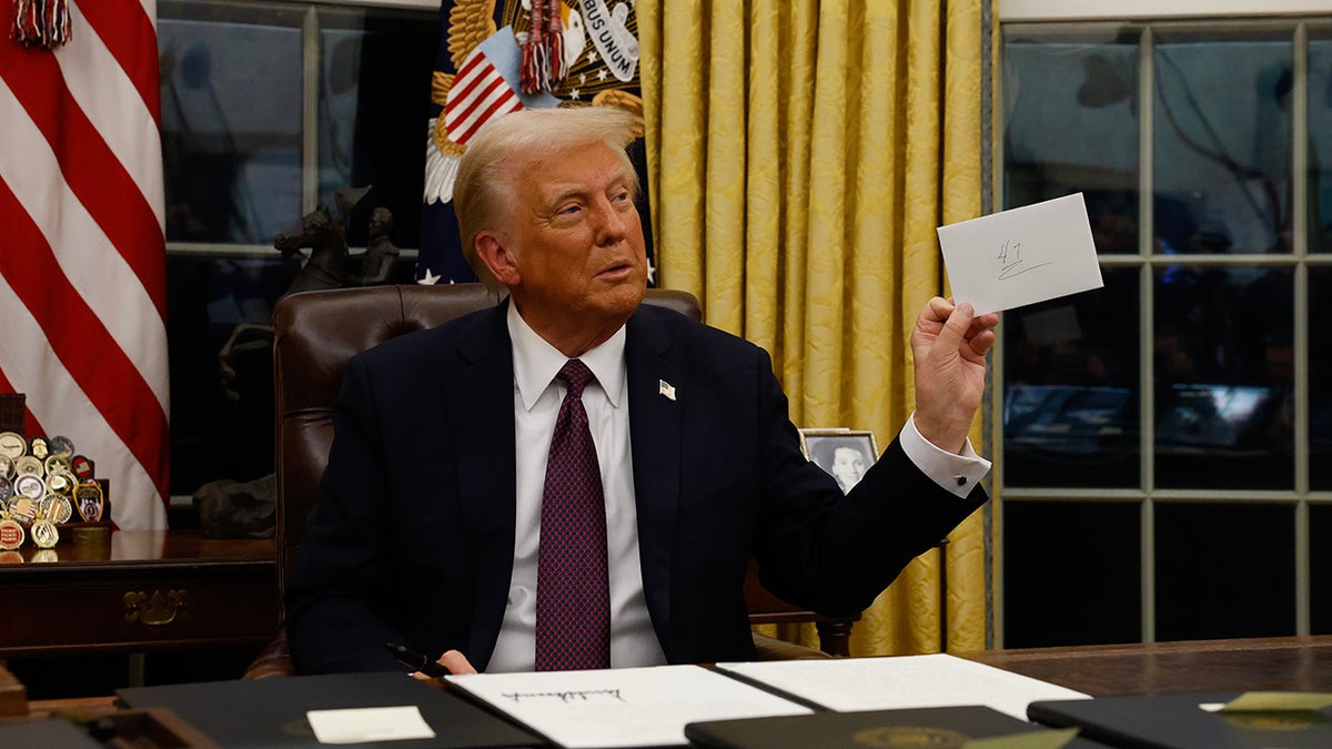 Donald Trump seated at desk in Oval Office holding envelope