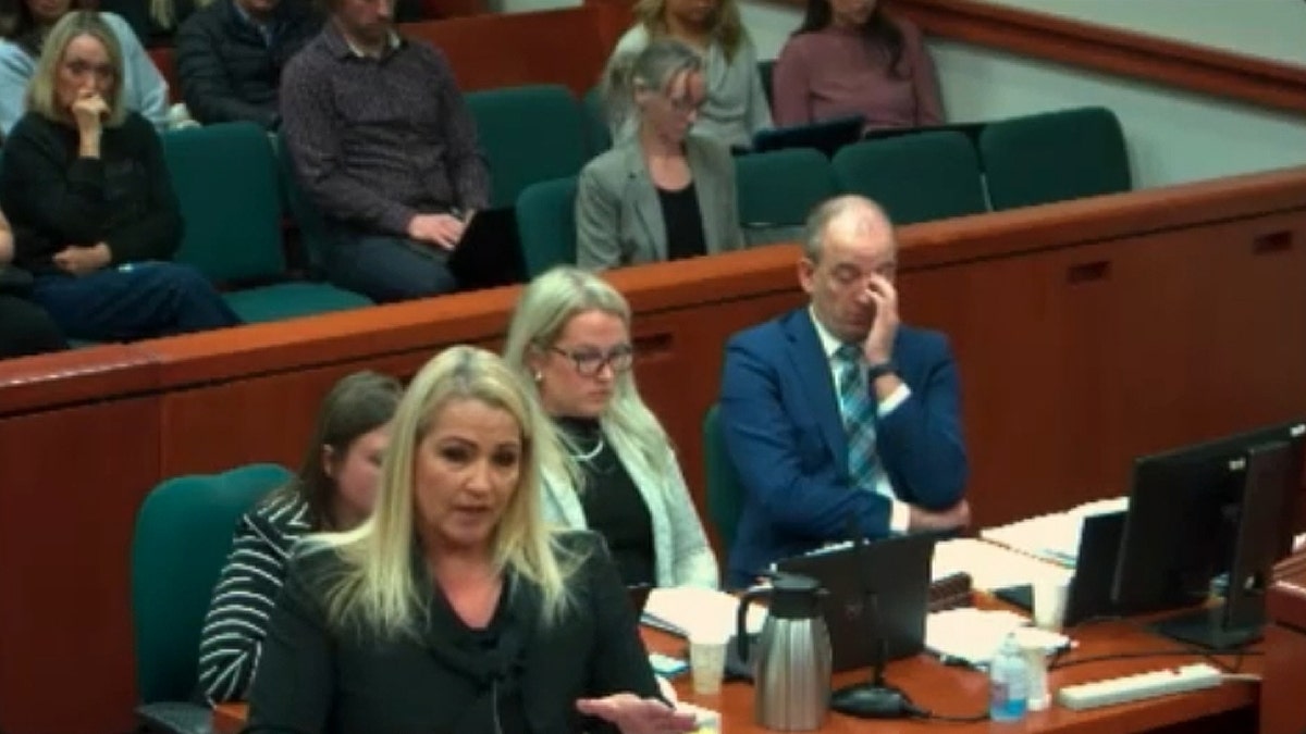 A woman in a black blouse speaks at a podium in a courtroom as others look on.