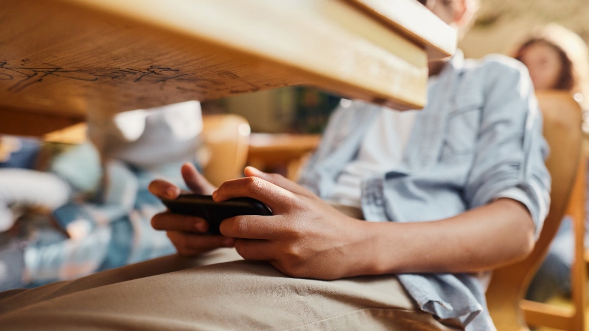 Close up of unrecognizable black student using cell phone under the table during a class at elementary school.