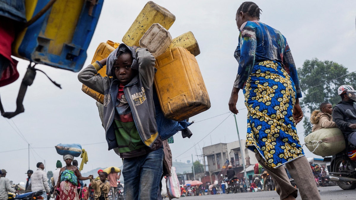 A child carries many plastic jugs slung on their back as a crowd flees fighting.