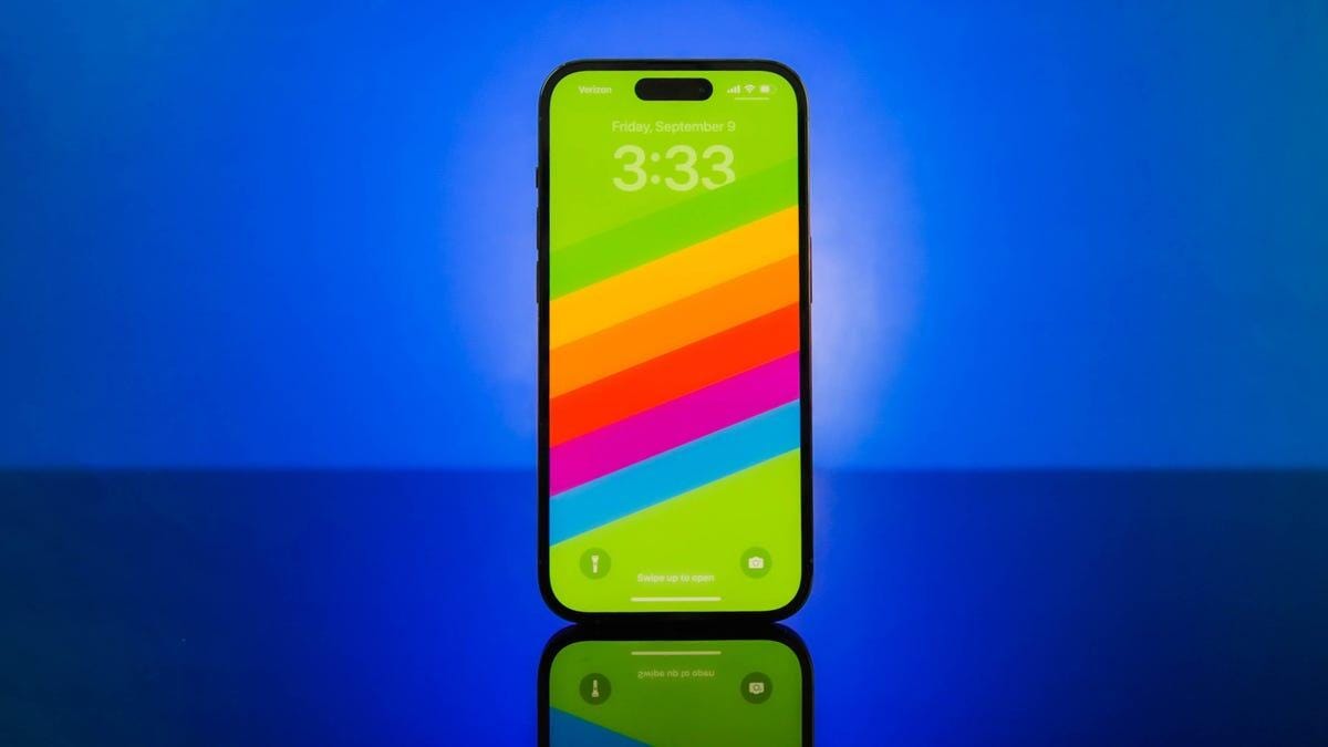 An iPhone 14 with a rainbow onscreen background against a blue backdrop