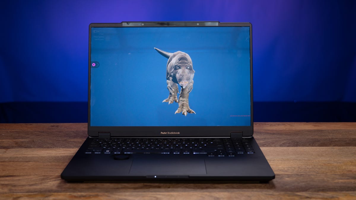 The StudioBook Pro 16 3D OLED sitting on a wood table and a blue background showing a dinosaur on the screen