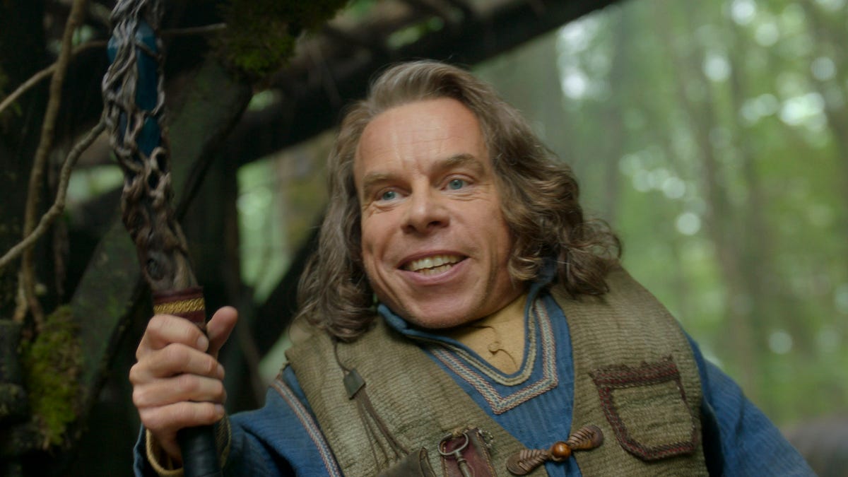 Actor Warwick Davis smiles and holds up a magic wand in a forest in fantasy TV series Willow.