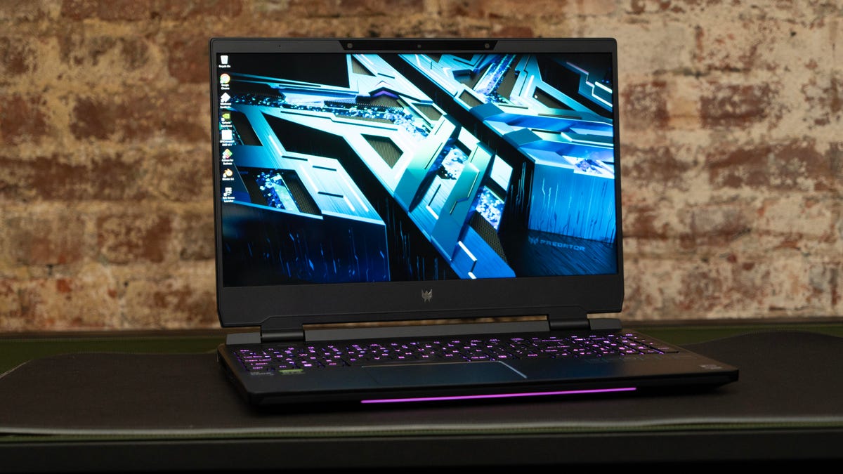 The Helios 300 sitting open on a desk against a brick wall with the blue geometric Windows wallpaper displayed.