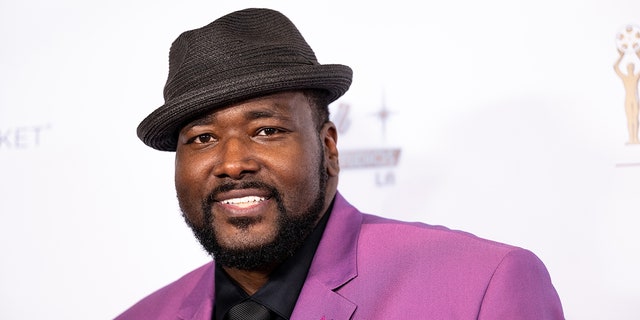 Quinton Aaron in a purple suit and black hat that is slightly tiled on the carpet