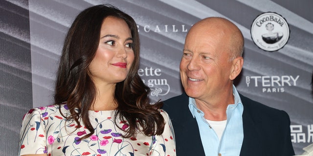 Emma Heming Willis in a white dress with flowers looks to her left as Bruce Willis in a blue shirt and navy blazer looks at her adoringly on the carpet