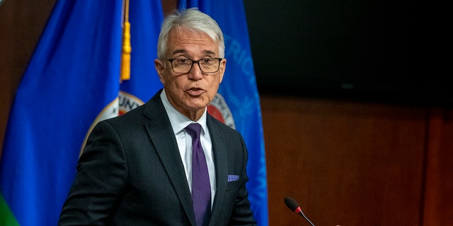 Gascon speaking in front of a microphone, wearing a suit and tie and a flag behind him