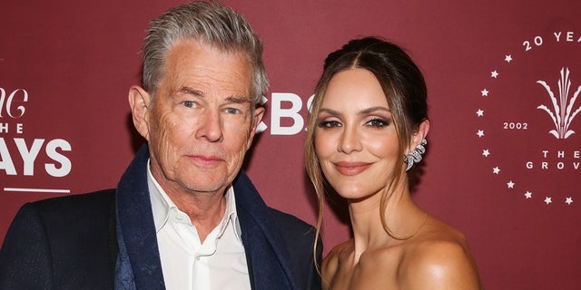 David Foster in a white shirt and dark jacket poses alongside his wife Katharine on the red carpet, she wears her hair in an up-do with two dangly pieces falling forward