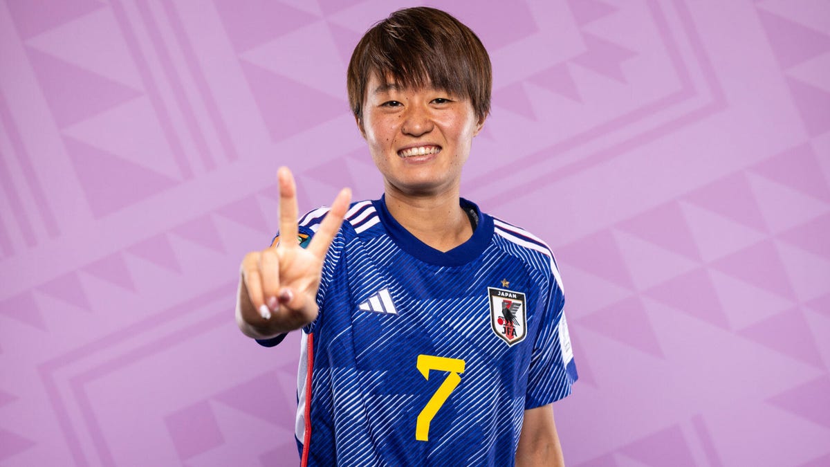 Japan soccer player Hinata Miyazawa gesturing a peace sign while smiling and standing in front of a purple and pink patterned background.