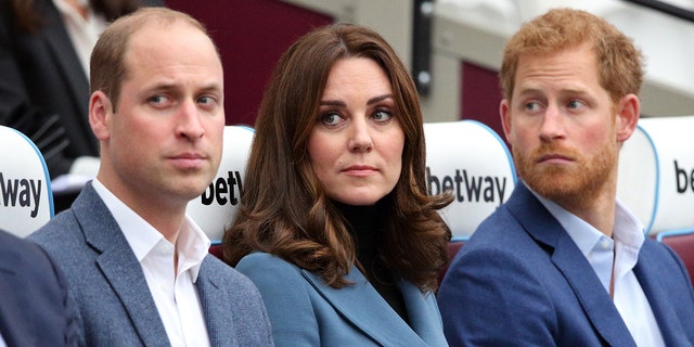 Kate Middleton wearing a blue blazer with a black sweater sitting in between Prince William and Prince Harry wearing various shades of blue and grey