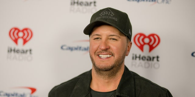 Luke Bryan in a black jean jacket and black hat on the carpet for iHeartCountry festival