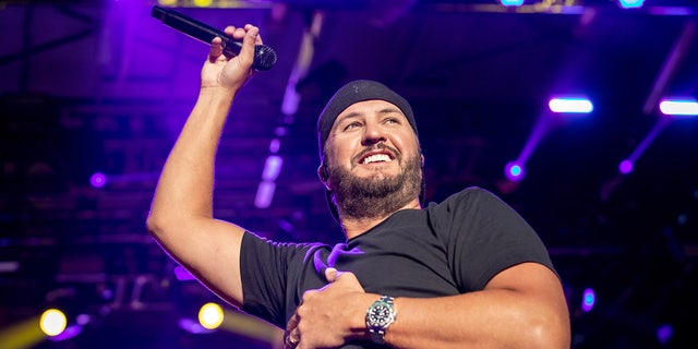 Luke Bryan in a black shirt and black backward hat raises his arm in the air over his head while on stage in Illinois