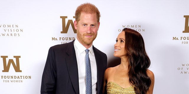 Prince Harry wearing a navy suit and a white shirt with a light blue tie posing next to Meghan Markle wearing a strapless gold dress