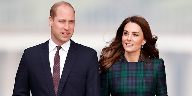 A close-up of Prince William wearing a dark blue blazer with a white shirt and red tie standing next to Kate Middleton in a green plaid dress