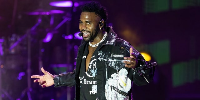 Jason Derulo in a ripped black shirt, black jacket that has been painted sings on stage