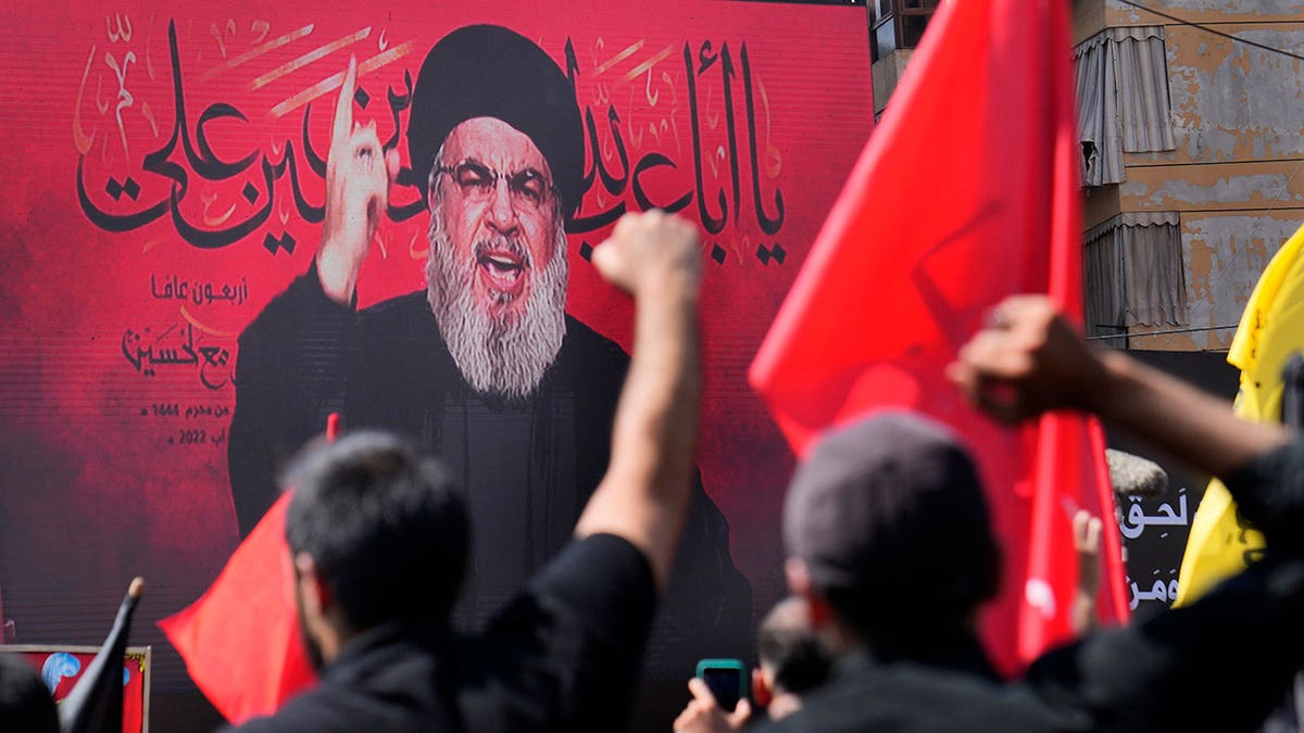 A mural of Hassan Nasrallah with a red background and people cheering in front of it