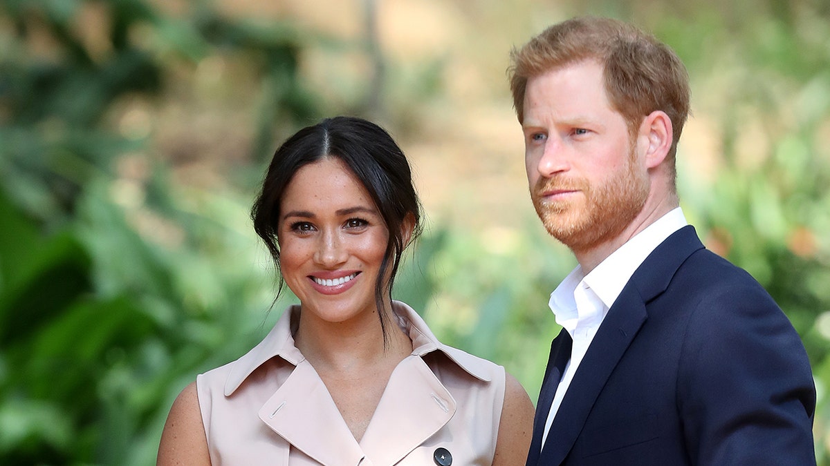 Meghan Markle smiling in a beige sleeveless dress while Prince Harry in a navy suit and white collared shirt looks serious.