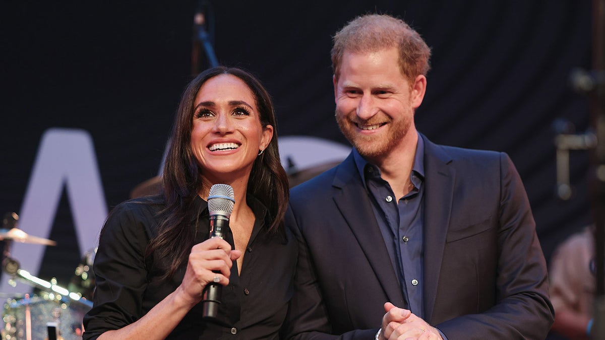 Meghan Markle wearing a black shirt and holding a mic next to Prince Harry wearing a dark blazer and a blue shirt.