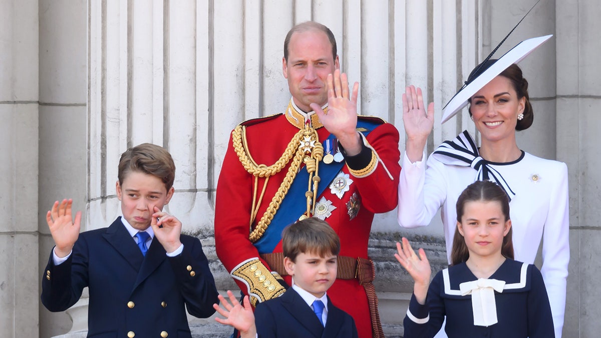 Prince William in red, along with Kate Middleton in white and their three children all wave from the balcony at Buckingham Palace