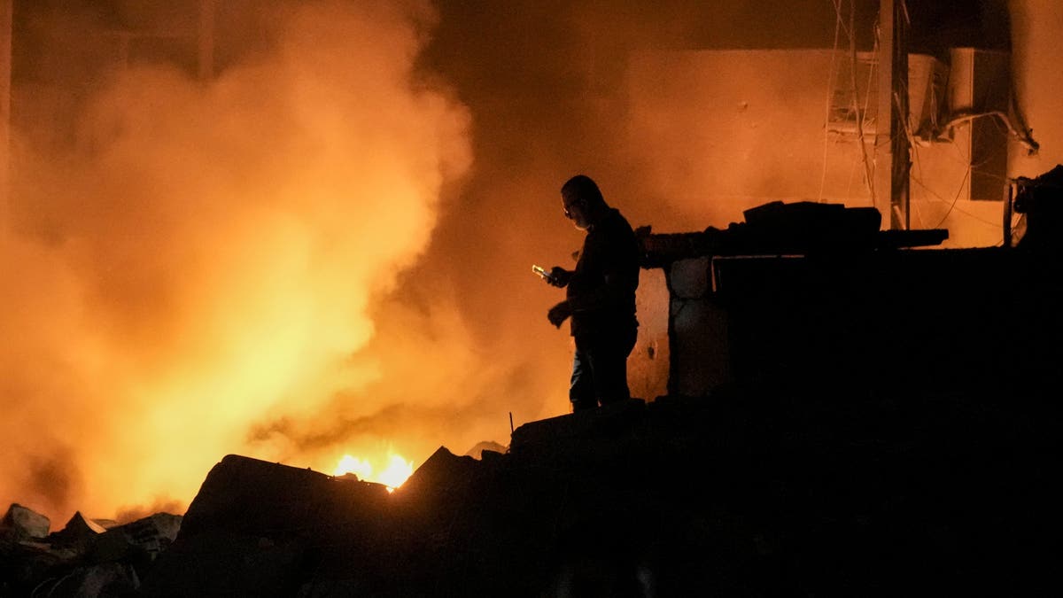 A man uses his mobile phone as flames and smoke rise at the scene of buildings hit by an Israeli airstrike in central Beirut, Lebanon, Thursday, Oct. 10, 2024. (AP Photo/Bilal Hussein)