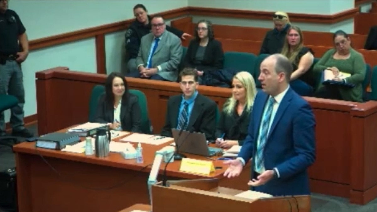 A man in a blue suit speaks at a podium in a courtroom as others look on.