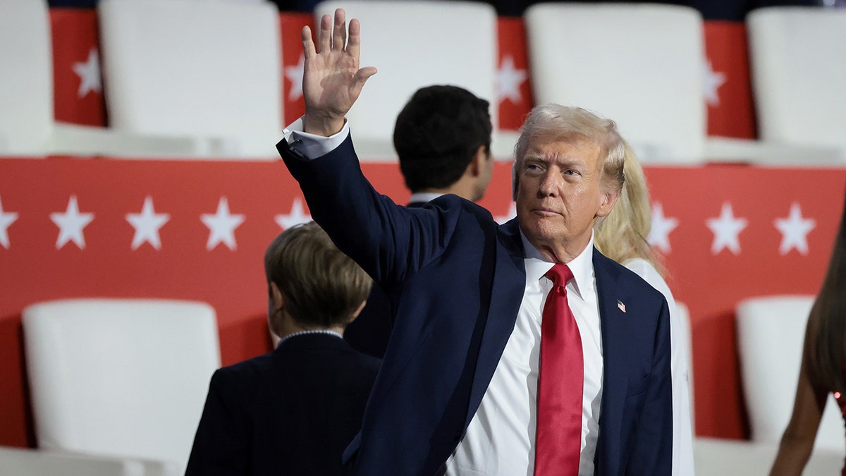 Donald Trump in a dark suit and red tie waves to the crowd and looks to his right